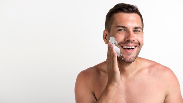 Portrait of young shirtless man applying shaving foam standing against white wall