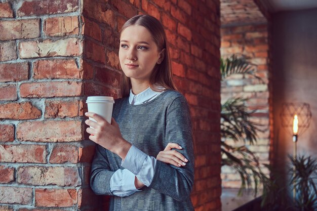 Portrait of a young slim sensual girl in a gray dress leaning against a brick wall with a morning coffee.