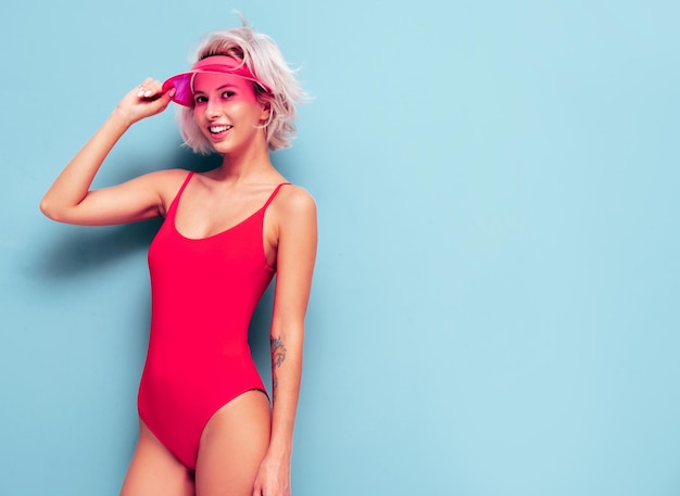 Free photo portrait of young smiling blond model in summer swimwear red bathing suit and transparent visor cap sexy carefree woman having fun and going crazy female posing near blue wall in studio