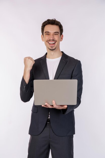 Portrait of young smiling businessman holding laptop in hands typing and browsing web pages while doing a winning closed fist gesture isolated on white background