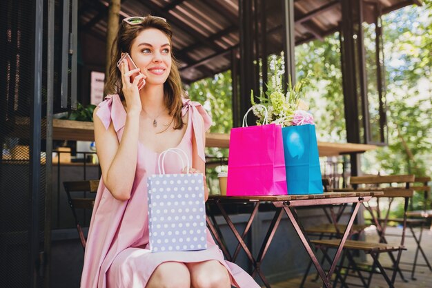 Portrait of young smiling happy attractive woman sitting in cafe talking on phone with shopping bags, summer fashion outfit, hipster style, pink cotton dress, trendy apparel