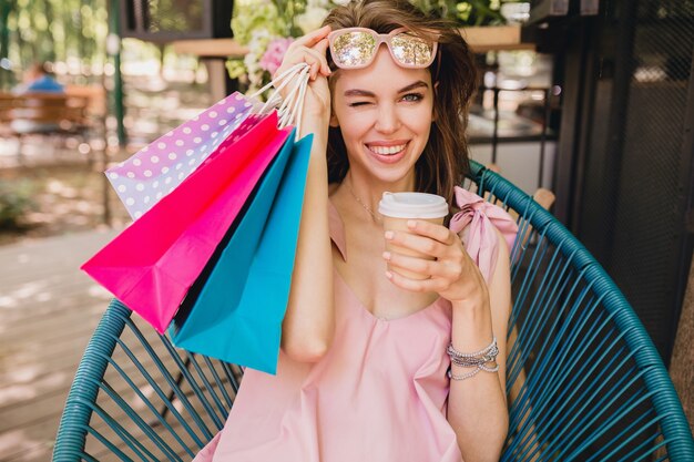 Portrait of young smiling happy pretty woman with excited face expression sitting in cafe with shopping bags drinking coffee, summer fashion outfit, hipster style, pink cotton dress, trendy apparel