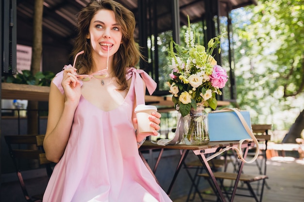 Portrait of young smiling happy pretty woman with sitting in cafe drinking coffee, summer fashion outfit, pink cotton dress, trendy apparel accessories