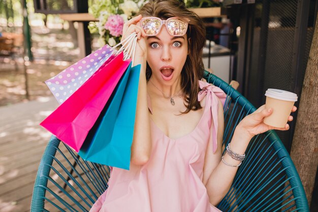 Portrait of young smiling happy pretty woman with surprised face expression sitting in cafe with shopping bags drinking coffee, summer fashion outfit, pink cotton dress, trendy apparel