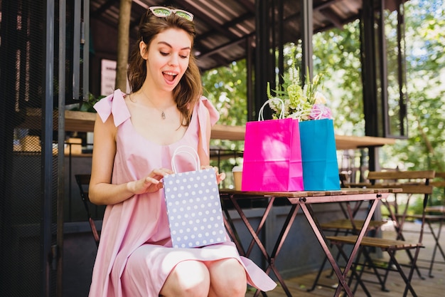 Portrait of young smiling happy pretty woman with surprised face expression sitting in cafe with shopping bags, summer fashion outfit, pink cotton dress, trendy apparel