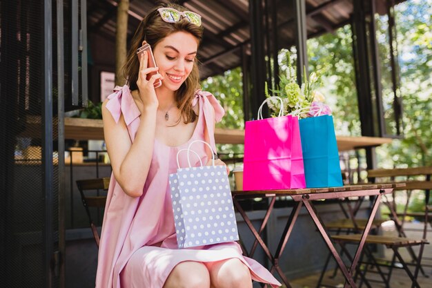 Portrait of young smiling happy pretty woman with surprised face expression sitting in cafe with shopping bags talking on phone, summer fashion outfit, pink cotton dress, trendy apparel