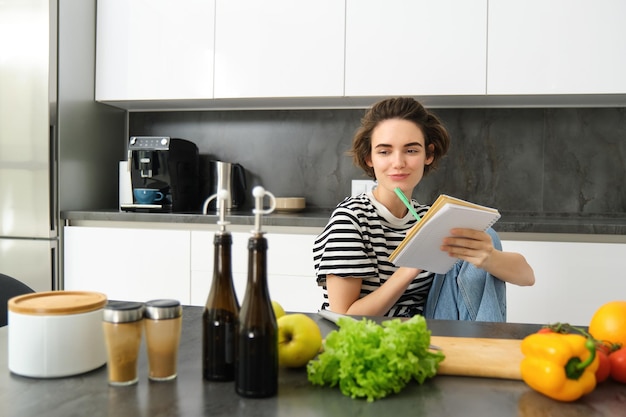 Free photo portrait of young smiling woman in kitchen holding notebook making notes for recipe writing grocery