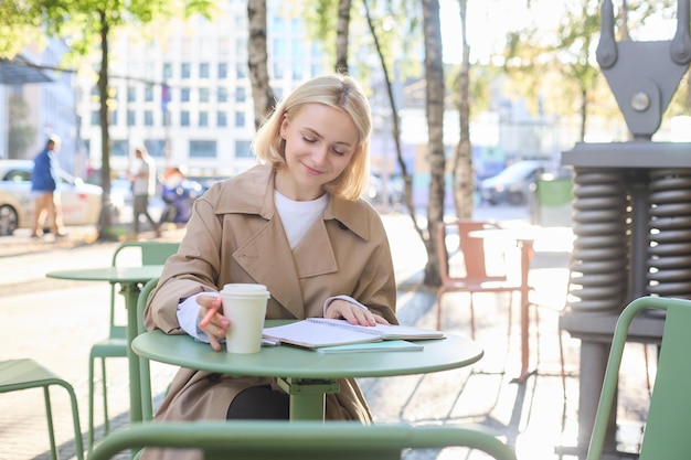 Free Photo portrait of young smiling woman working outdoors sitting with notebook in cafe and writing drinking