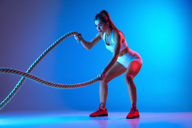 Portrait of young spotive girl doing exercises with rope keeping body fit isolated over blue background in neon