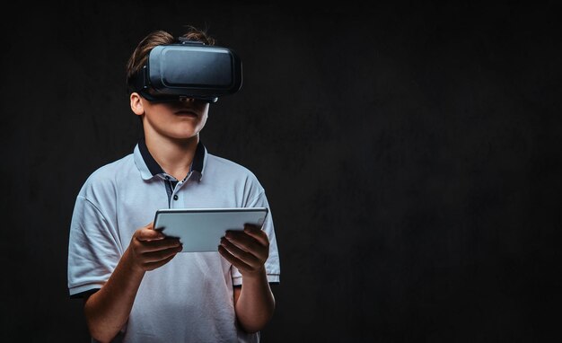 Portrait of a young student boy dressed in a white t-shirt using virtual reality glasses and tablet computer. Isolated on a dark background.