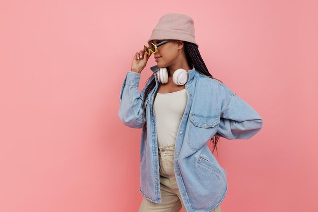 Portrait of young stylish girl with white headphones in trendy sunglasses posing isolated over pink studio background