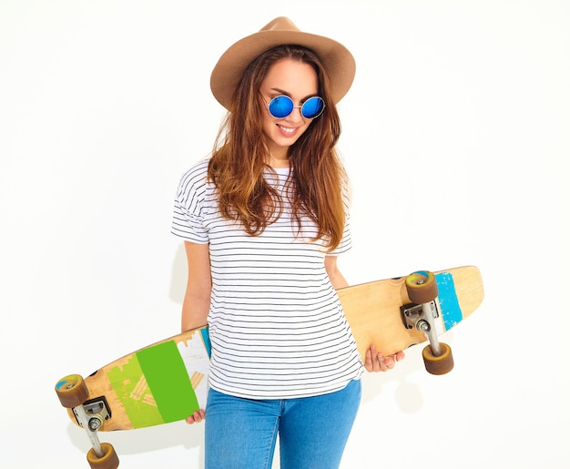 Portrait of young stylish woman model in casual summer clothes in brown hat posing with longboard desk. Isolated on white