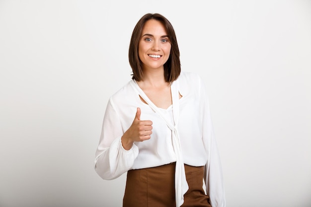 Free Photo portrait of young successful business woman smiling, showing thumb up, looking at front, over white wall.