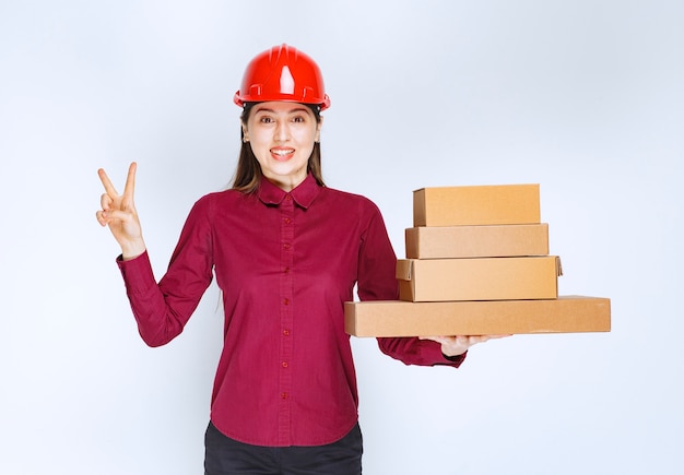 Portrait of a young woman in crash helmet with paper boxes showing victory sign . 