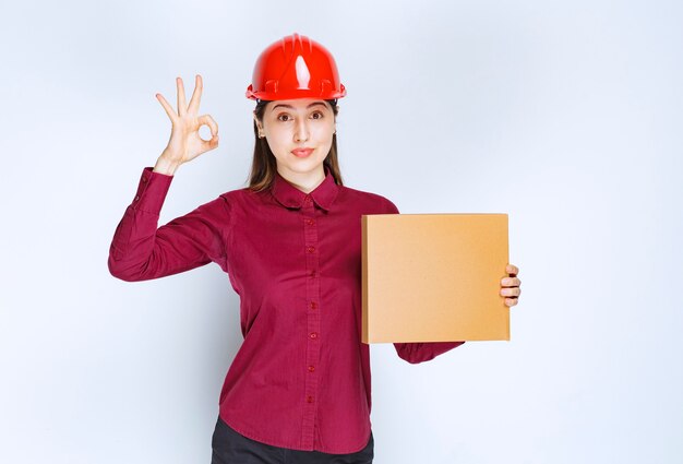 Portrait of a young woman in crash helmet with small paper box showing ok gesture . 