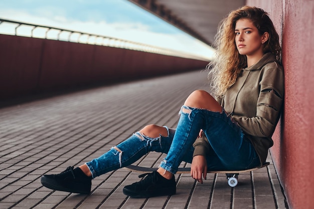 Free photo portrait of a young woman dressed in a hoodie and ripped jeans leaning on a wall while sitting on a skateboard at bridge footway.