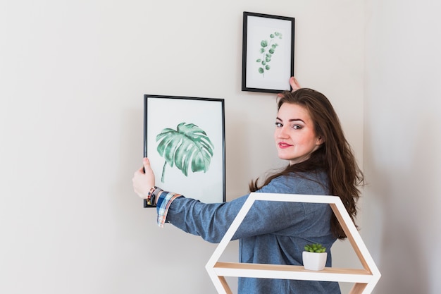 Portrait of a young woman fixing the picture frame on wall looking to camera