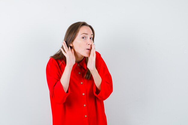 Portrait of young woman keeping hand near mouth while overhearing secret in red blouse and looking curious front view