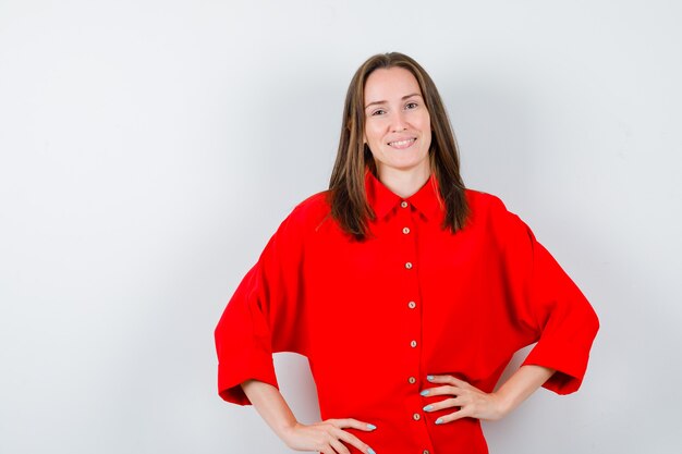 Portrait of young woman keeping hands on waist in red blouse and looking jovial front view