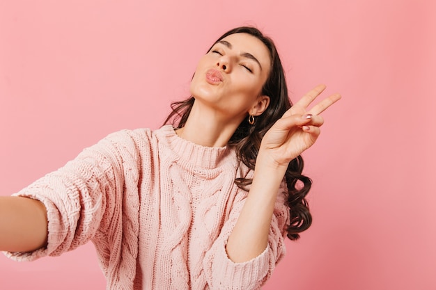 Portrait of young woman in knitted outfit taking selfie in pink studio. Lady with closed eyes sends air kiss and shows peace sign.