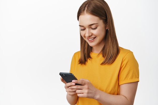 Free photo portrait of young woman look at smartphone screen and smiling, reading message on cell phone, using mobile app or messanger, standing against white background.