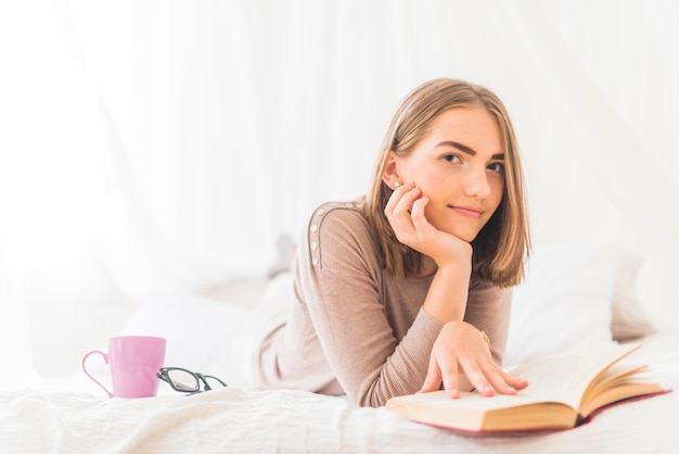 Portrait of young woman lying on bed for reading book with coffee