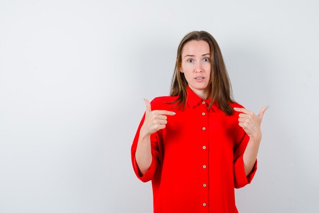 Portrait of young woman pointing at herself in red blouse and looking puzzled front view