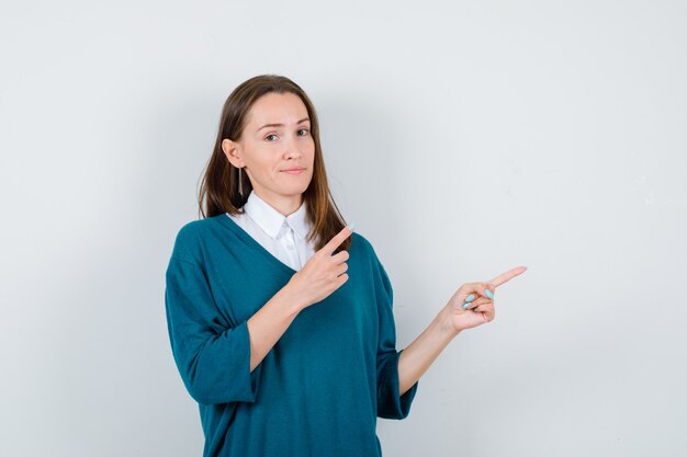 Portrait of young woman pointing at upper right corner in sweater over white shirt and looking confident front view