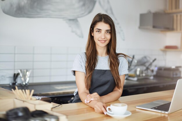 Portrait of young woman running a successful cafe drinking freshly made capuccino and using a laptop