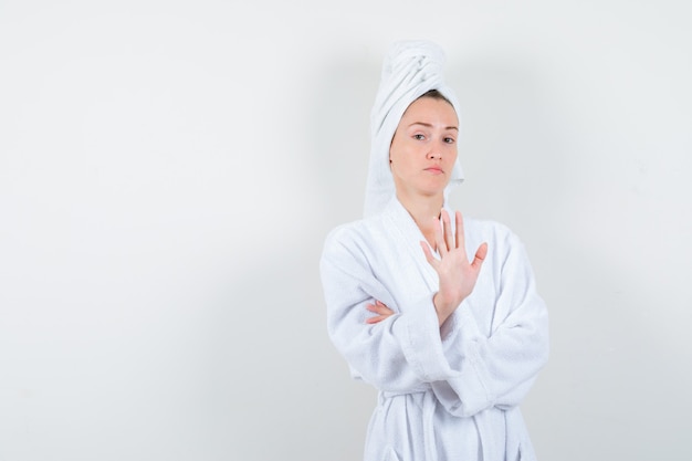 Portrait of young woman showing stop gesture in white bathrobe, towel and looking confident front view