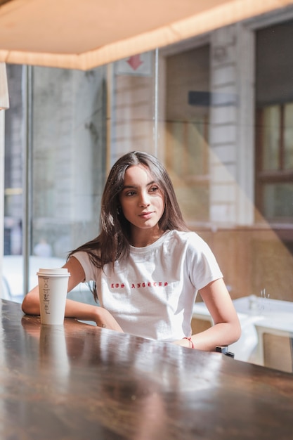 Free Photo portrait of a young woman sitting in cafe with disposable coffee cup on table