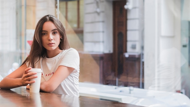 Portrait of a young woman sitting in cafe with takeaway coffee cup in hand looking at camera