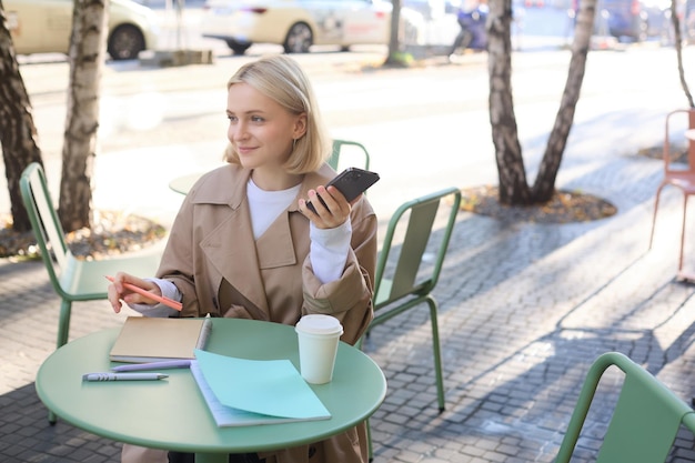 Free Photo portrait of young woman studying sitting in outdoor cafe with smartphone and journals doing homework