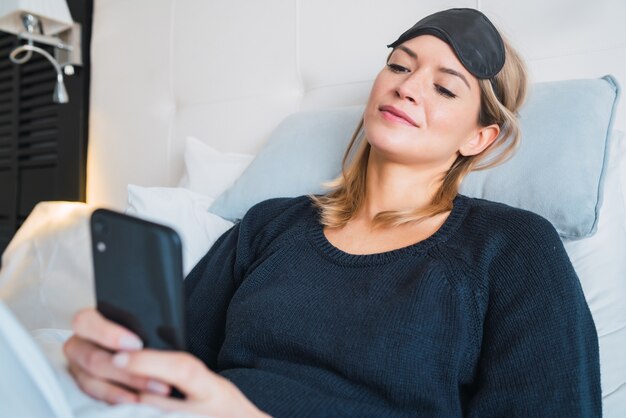 Portrait of young woman using her mobile phone while laying on bed at hotel room. Travel and lifestyle concept.