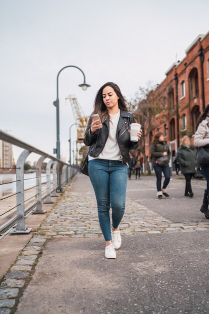 Portrait of young woman using her mobile phone while walking with cup of coffee. Urban and communication concept.