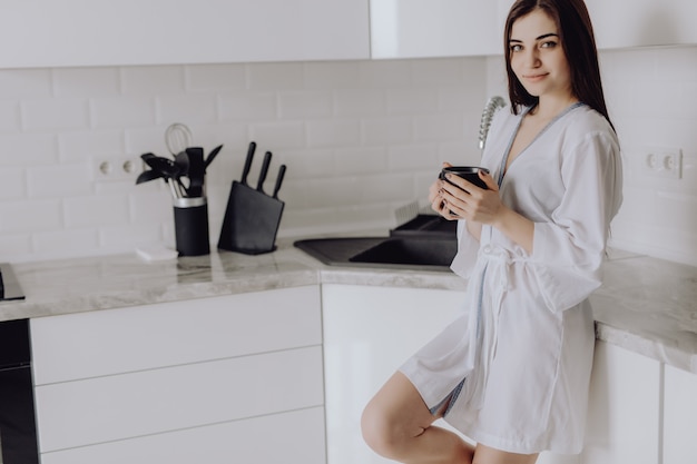 Portrait of young woman in white robe after bath holds a cup with coffee or tea against kitchen wall.