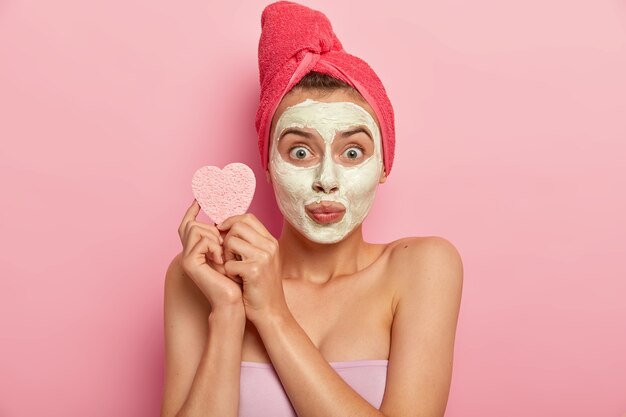 Portrait of young woman with mineral natural mask on face, has daily skin care treatment, holds cosmetic sponge for wiping complexion, purifies skin and removes dead skin cell, has bath at home