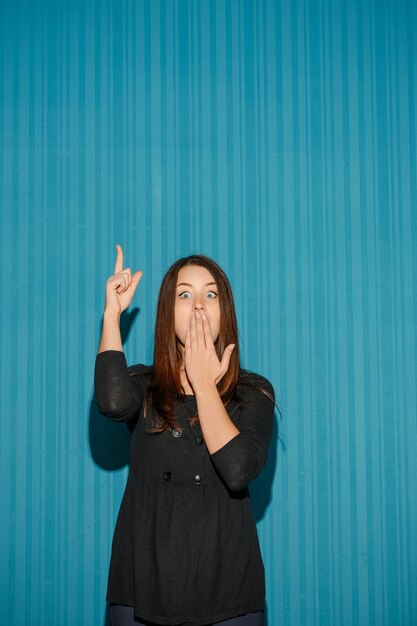 Portrait of young woman with shocked facial expression over blue studio background pointing up