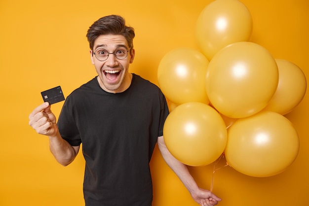 Positive adult man holds credit card spends money on party and celebration glad to celebrate anniversary in company of friends wears casual clothes poses with balloons against yellow backround