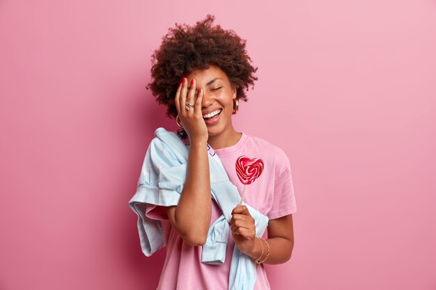 Positive African American teenage girl makes face palm, has fun, dressed in casual clothes, holds heart shaped lollipop, enjoys spare time, poses against pink wall. Confectionery concept