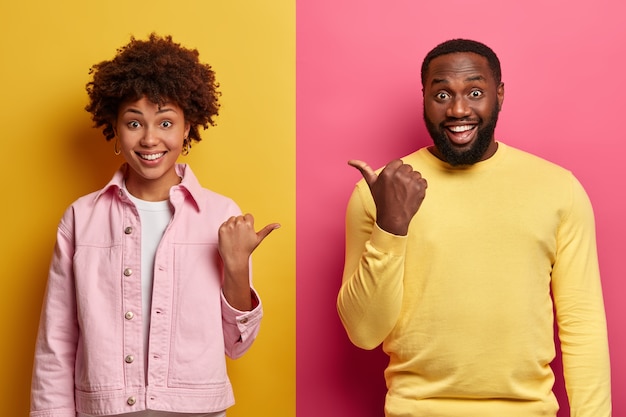 Free photo positive african american woman and man point thumb at each other, smile and look enthusiastic, dressed in casual clothes, isolated on pink and yellow wall, have happy mood, friendly relations