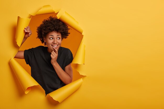 Positive Afro girl holds chin, focused aside, feels winsome and cheerful, wears casual t shirt, poses in paper hole alone