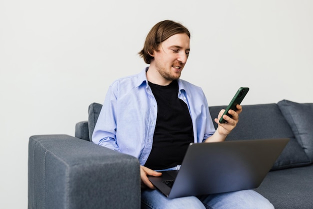 Positive bearded man with laptop talking on smartphone sitting on sofa in living room