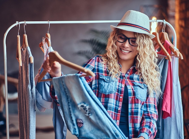 Free photo positive blonde female in black eyeglasses chooses fashionable clothes on the coat rack.