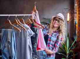 Free photo positive blonde female in black eyeglasses chooses fashionable clothes on the coat rack.