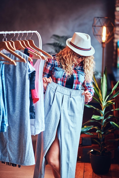 Positive blonde female in black eyeglasses chooses fashionable clothes on the coat rack.