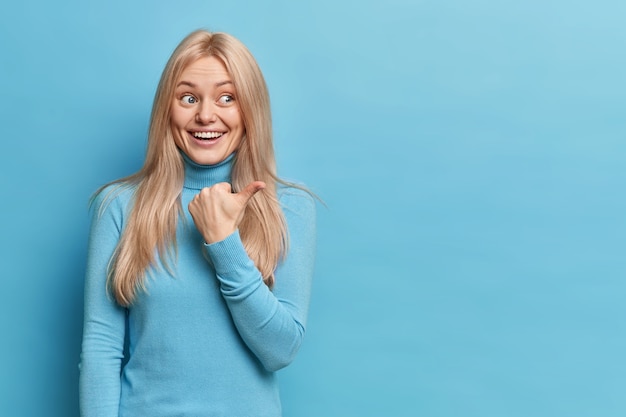 Positive blonde smiling young European woman points thumb away at blank copy space