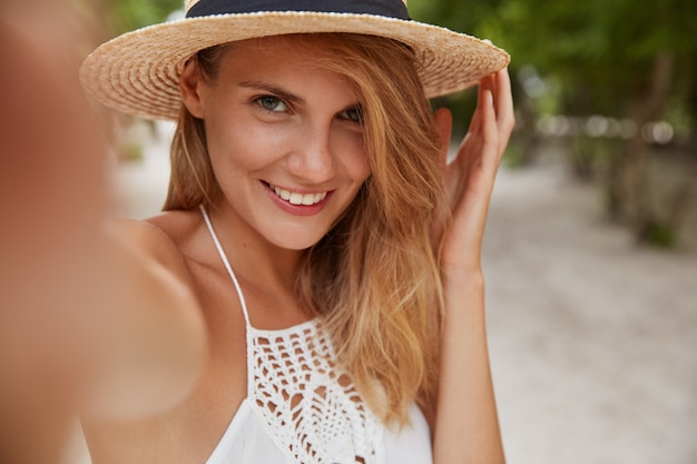 Positive blonde young female with cheerful expression makes selfie as poses outdoor on tropical island, wears fashionable summer hat and white dress