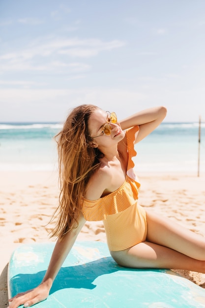 Positive brown-haired caucasian girl posing at beach with happy smile. Outdoor portrait of amazing brunette woman in orange swimwear relaxing after surfing in ocean.