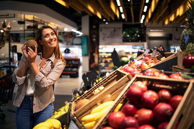 Positive brunette woman holding coconut at grocery store fruit department